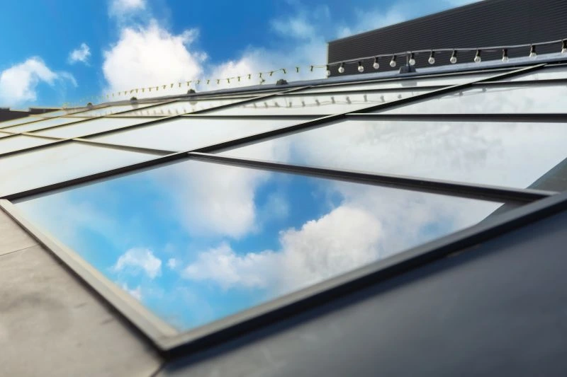Low-angle view of glass skylights reflecting a blue sky with white clouds on a roof.